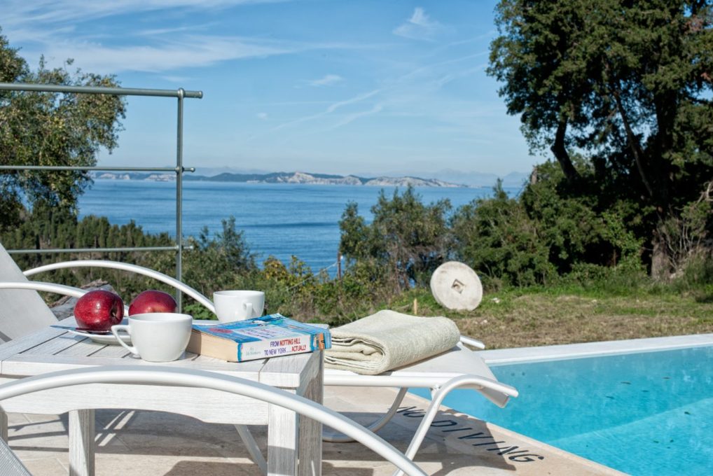 Coffee, fruit and a book on a poolside table at Mistral House with the sea beyond