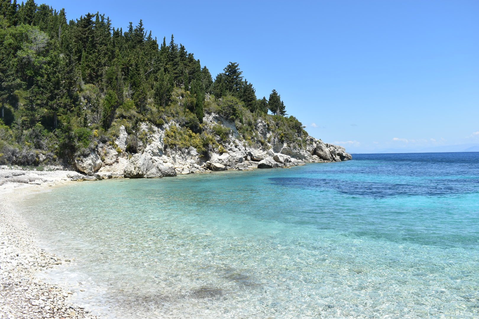 A quiet pebble cove on Paxos with clear turquoise water and pine-covered headland